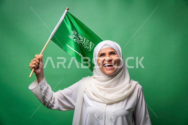 الاحتفال بالأعياد والمناسبات الوطنية، استذكار اليوم الوطني السعودي 23 سبتمبر، رفع راية التوحيد والإسلام، الفخر و الاعتزاز بالانتماء للوطن، يوم العلم 11 مارس، بورتريه مقرب لامرأة مسنة عربية خليجية سعودية محجبة تحمل بيدها علم السعودية الاخضر، خلفية خضراء، كروما