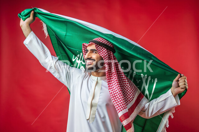 Flag Day March 11, Celebrating National Holidays and Occasions, Remembering Saudi National Day September 23, Raising the Banner of Monotheism and Islam, Pride and Honor of Belonging to the Nation, Close-up Portrait of a Saudi Arabian Gulf Man Holding the Green Saudi Flag, Red Background