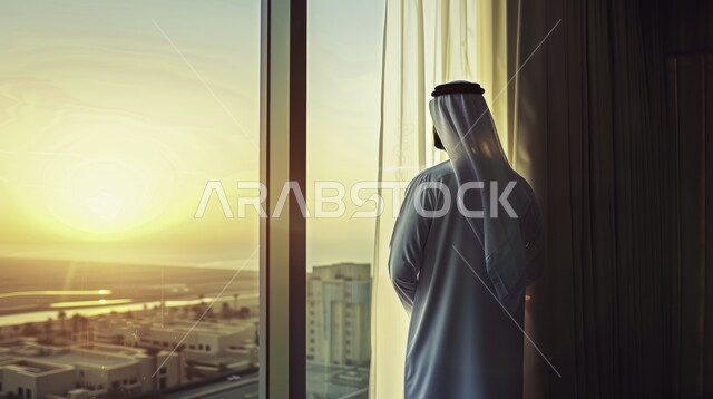  تقدم وتطور الفن الهندسي المعماري في السعودية، التأمل وشرود الذهن، التفكير في شيء ما، صورة مقربة من الخلف لرجل عربي خليجي سعودي يرتدي الشماغ والثوب السعودي التقليدي يقف امام واجهه زجاجية كبيرة، الاستمتاع بالنظر الى العالم الخارجي من خلال النافذة
