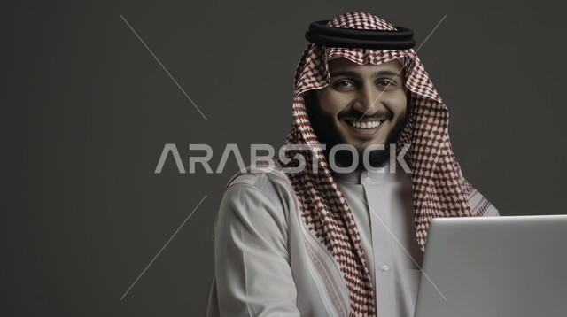 النظر إلى الكاميرا بإيماءات البهجة والسرور، مهن ووظائف سعودية مكتبية، استخدام الأجهزة والتقنيات الحديثة لتسهيل الأعمال, بورتريه مقرب لرجل أعمال عربي سعودي خليجي يرتدي الزي التقليدي يجلس على مكتب أمام الحاسوب المحمول, إجراء مقابلات عمل أونلاين, خلفية رمادية