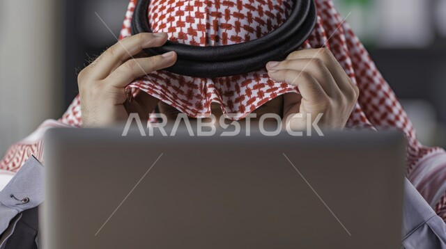 مفهوم ضغط العمل, إيماءات الصداع والضجر, مهن ووظائف مكتبية, صورة مقربة لرجل عربي خليجي سعودي يرتدي الشماغ والثوب التقليدي يضع يديه على رأسه يقوم باستخدام الحاسب المحمول, تعابير الحزن والتعب, إدارة وتنظيم الأعمال ضمن مقر الشركة