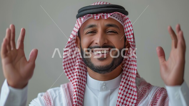 تعابير الترحيب والاستقبال، الشعور بالفرح والسرور، بورتريه مقرب لرجل عربي خليجي سعودي يرتدي الثوب التقليدي والشماغ يرفع يديه بإيماءات السعادة، الاهتمام بالأناقة والمظهر الخارجي، خلفية رمادية 