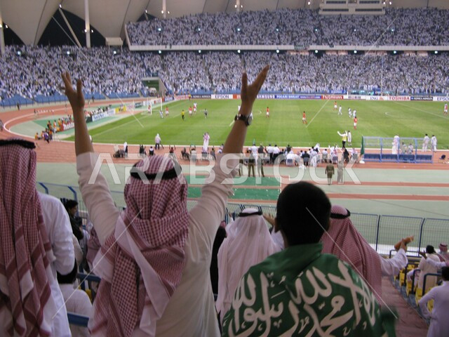 الشغف بكرة القدم، رفع اليدين بإيماءات السعادة والفرح بالفوز، صورة من الخلف لشاب عربي خليجي سعودي يرتدي الثوب التقليدي والشماغ يجلس في الاستاد الرياضي, ولد من الجماهير يلبس العلم السعودي, تشجيع فريق المملكة العربية السعودية في نهائيات كأس العالم