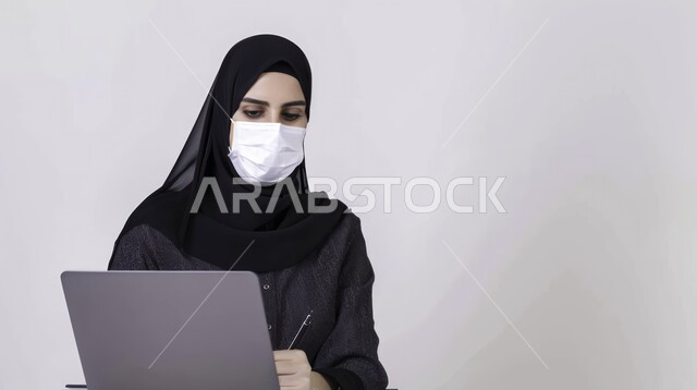 بيئة عمل مريحة، استخدام التقنيات والاجهزة الحديثة في إنجاز المهام المكتبية، بورتريه مقرب لامرأة عربية خليجية سعودية ترتدي العباية والحجاب والكمامة تقوم بإستخدام جهاز الحاسوب المحمول، مهن ووظائف نسائية في المملكة، خلفية بيضاء