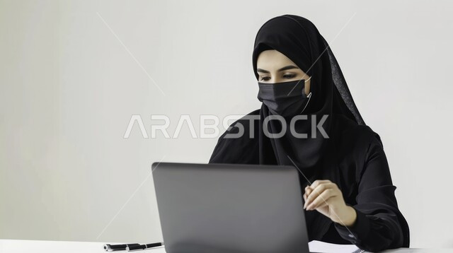 استخدام التقنيات والاجهزة الحديثة في إنجاز المهام المكتبية، بورتريه مقرب لامرأة عربية خليجية سعودية ترتدي العباية والحجاب وكمامة سوداء تقوم بإستخدام جهاز الحاسوب المحمول، مهن ووظائف نسائية في المملكة، بيئة عمل مريحة، خلفية بيضاء
