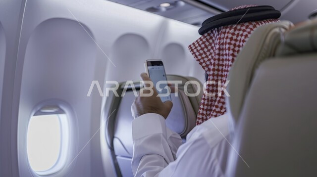 Saudi Airlines, tourist flights, making a video call via mobile phone, the concept of tourism and travel, a close-up picture from the side of a Saudi Gulf Arab man wearing the traditional thobe and shemagh sitting in the plane seat holding a mobile phone in his hand, using modern devices and technologies