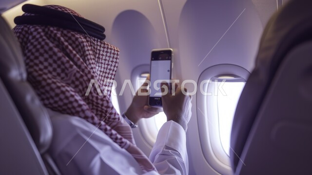 Using modern devices and technologies, Saudi Airlines, tourist flights, making a video call via mobile phone, the concept of tourism and travel, a close-up picture from the side of a Saudi Gulf Arab man wearing the traditional thobe and shemagh, sitting in the plane seat, holding a mobile phone in his hand.