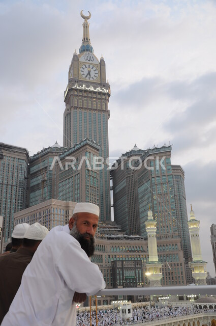 مقاصد واماكن لأداء العبادات والتقرب الى الله تعالى، معالم دينية اسلامية مقدسة في المملكة العربية السعودية، الفن الهندسي المعماري لبناء الابراج وناطحات السحاب، صورة مقربة لرجل يقف امام مبنى برج الساعة الملكي يرتدي الزي الابيض في مكة المكرمة