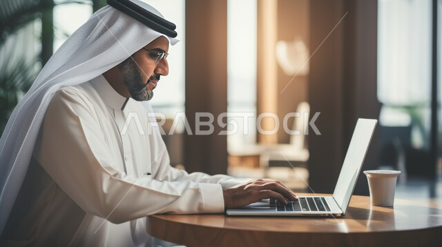 Completing the required tasks via the laptop, the field of economics, finance and business, Saudi office jobs and professions, integrating work with technology, managing and organizing the company’s affairs through technical devices, an Arab Gulf Emirati man wearing the kandora and ghutra uniform working in the office
