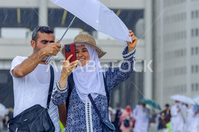 توثيق اللحظات السعيدة في مكة المكرمة , زيارة بيت الله الحرام , زوجان مسلمان يرتديان الزي الكاجوال يحملان بيدهما شمسية بيضاء يقومان بالتقاط صورة سيلفي تذكارية , إيماءات الفرح لأداء فريضة الحج و العمرة , أجواء شتوية في المملكة العربية السعودية