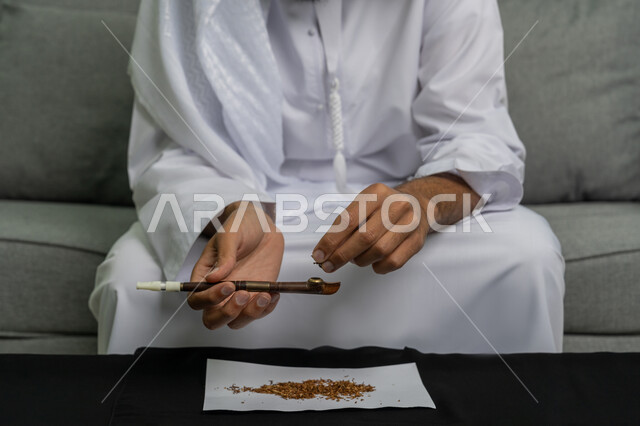 الأثار السلبية للادمان على التبغ والمخدرات، القيام بعادات سلوكية ضارة وتعاطي المواد السامة، المعاناة من الاضطرابات النفسية والمشاكل الصحية، صورة مقربة لأيدي شاب عربي خليجي إماراتي يرتدي الكندورة والغترة البيضاء يجلس على اريكة يقوم بتعبئة التبغ داخل سيجار معدني