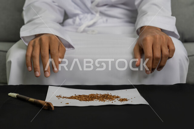 القيام بعادات سلوكية ضارة وتعاطي المواد السامة، الأثار السلبية للادمان على التبغ والمخدرات، المعاناة من الاضطرابات النفسية والمشاكل الصحية، صورة مقربة لأيدي شاب عربي خليجي إماراتي يرتدي الكندورة والغترة البيضاء يجلسةأمام طاولة تحوي سيجار معدني ومجموعة من التبغ