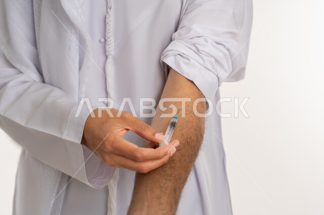 تعاطي المخدرات عن طريق الحقن في الأوردة، القيام بعادات سلوكية ضارة وتعاطي المواد السامة، المعاناة من الاضطرابات النفسية والمشاكل الصحية، بورتريه مقرب لأيدي شاب عربي خليجي إماراتي يرتدي الكندورة يقوم بأخذ ابرة مخدرة، خلفية بيضاء