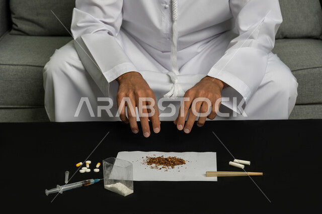 القيام بعادات سلوكية ضارة وتعاطي المواد السامة، الأثار السلبية للادمان على التبغ والمخدرات، المعاناة من الاضطرابات النفسية والمشاكل الصحية، صورة مقربة لأيدي شاب عربي خليجي إماراتي يرتدي الكندورة والغترة البيضاء يجلس على اريكة يقوم بلف عقب السجائر