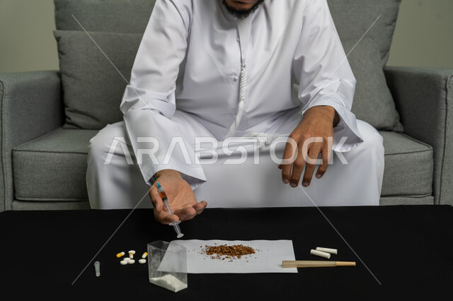 المعاناة من الاضطرابات النفسية والمشاكل الصحية، تعاطي المخدرات عن طريق الحقن في الأوردة، القيام بعادات سلوكية ضارة وتعاطي المواد السامة، صورة مقربة لأيدي شاب عربي خليجي إماراتي يرتدي الكندورة يقوم بأخذ ابرة مخدرة