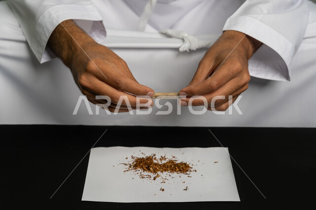 الأثار السلبية للادمان على التبغ والمخدرات، القيام بعادات سلوكية ضارة وتعاطي المواد السامة، المعاناة من الاضطرابات النفسية والمشاكل الصحية، صورة مقربة لأيدي شاب عربي خليجي إماراتي يرتدي الكندورة والغترة البيضاء يجلس على اريكة يقوم بلف عقب السجائر