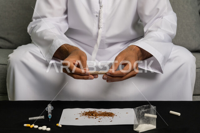 الأثار السلبية للادمان على التبغ والمخدرات، القيام بعادات سلوكية ضارة وتعاطي المواد السامة، المعاناة من الاضطرابات النفسية والمشاكل الصحية، صورة مقربة لرجل عربي خليجي إماراتي يرتدي الكندورة والغترة البيضاء يجلس على اريكة يقوم بلف عقب السجائر