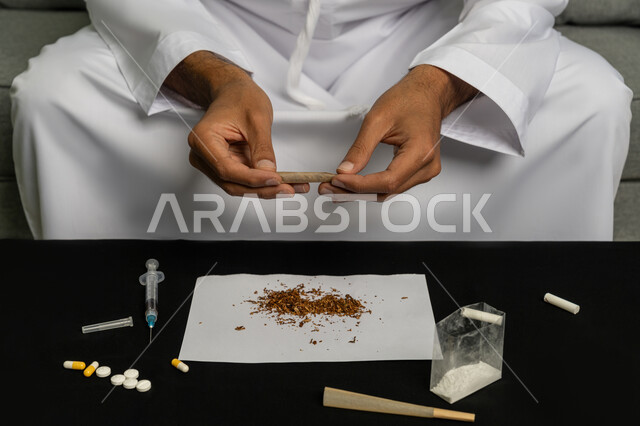 القيام بعادات سلوكية ضارة وتعاطي المواد السامة، الأثار السلبية للادمان على التبغ والمخدرات، المعاناة من الاضطرابات النفسية والمشاكل الصحية، صورة مقربة لرجل عربي خليجي إماراتي يرتدي الكندورة والغترة البيضاء يجلس على اريكة يقوم بلف عقب السجائر