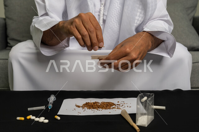 المعاناة من الاضطرابات النفسية والمشاكل الصحية، القيام بعادات سلوكية ضارة وتعاطي المواد السامة، الأثار السلبية للادمان على التبغ والمخدرات، صورة مقربة لرجل عربي خليجي إماراتي يرتدي الكندورة والغترة البيضاء يجلس على اريكة يقوم بلف عقب السجائر