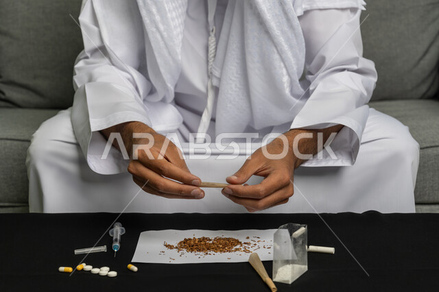 القيام بعادات سلوكية ضارة وتعاطي المواد السامة، الأثار السلبية للادمان على التبغ والمخدرات، المعاناة من الاضطرابات النفسية والمشاكل الصحية، صورة مقربة لرجل عربي خليجي إماراتي يرتدي الكندورة والغترة البيضاء يجلس على اريكة يقوم بلف عقب السجائر