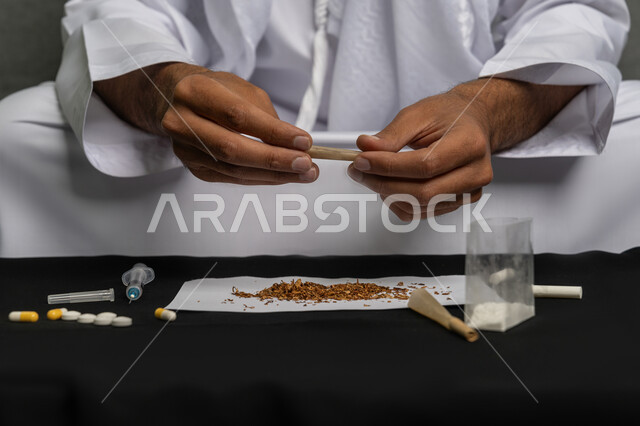 الأثار السلبية للادمان على التبغ والمخدرات، القيام بعادات سلوكية ضارة وتعاطي المواد السامة، المعاناة من الاضطرابات النفسية والمشاكل الصحية، صورة مقربة لرجل عربي خليجي إماراتي يرتدي الكندورة والغترة البيضاء يجلس على اريكة يقوم بلف عقب السجائر