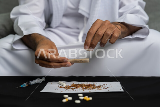 الأثار السلبية للادمان على التبغ والمخدرات، القيام بعادات سلوكية ضارة وتعاطي المواد السامة، المعاناة من الاضطرابات النفسية والمشاكل الصحية، صورة مقربة لرجل عربي خليجي إماراتي يرتدي الكندورة والغترة البيضاء يجلس على اريكة يقوم بلف عقب السجائر