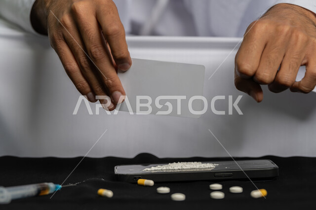 المعاناة من الاضطرابات النفسية والمشاكل الصحية، الآثار السلبية للادمان، القيام بعادات سلوكية ضارة، تعاطي المواد السامة، صورة مقربة لأيدي شاب عربي خليجي إماراتي يرتدي الكندورة والغترة يقوم بتحضير البودرة البيضاء لاستنشاقها