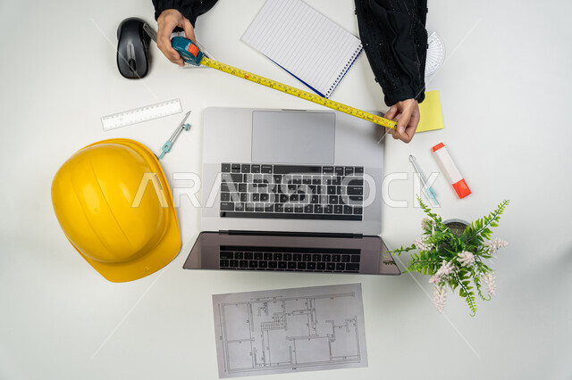 Working in the engineering sector, studying the basics of the construction plan, following up and managing the project, presenting and reviewing project manuscripts, vertical top portrait of an Arab Gulf Emirati female engineer working on a laptop, a Saudi woman in front of a desk with a safety helmet and plans, white background
