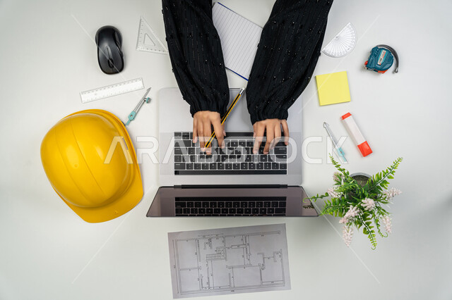 دراسة أساسيات المحطط الإنشائي, متابعة وإدارة المشروع, عرض ومراجعة مخطوطات المشاريع, العمل في القطاع الهندسي, بورتريه علوي عامودي ليدي مهندسة عربية خليجية اماراتية تعمل على الحاسوب المحمول، امرأة سعودية امام مكتب عليه خوذة الحماية والمخططات، خلفية بيضاء
