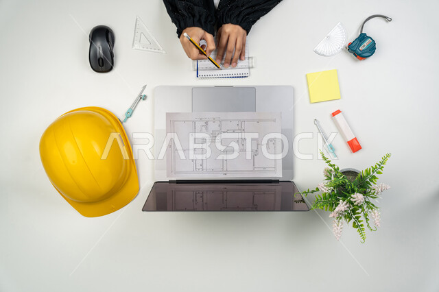 إدارة ومتابعة المشروع, عرض ومراجعة مخطوطات المشاريع, العمل في القطاع الهندسي, دراسة أساسيات المحطط الإنشائي, بورتريه علوي عامودي ليدي مهندسة عربية خليجية اماراتية تعمل على الحاسوب المحمول، امرأة سعودية امام مكتب عليه خوذة الحماية والمخططات، خلفية بيضاء