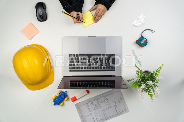 مراجعة وعرض مخطوطات المشاريع, العمل في القطاع الهندسي, دراسة أساسيات المحطط الإنشائي, بورتريه علوي عامودي ليدي مهندسة عربية خليجية اماراتية تعمل على الحاسوب المحمول، امرأة سعودية امام مكتب عليه خوذة الحماية والمخططات، إدارة ومتابعة المشروع, خلفية بيضاء