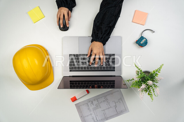 Working in the engineering sector, studying the basics of construction planning, vertical overhead portrait of an Emirati Gulf Arab female engineer working on a laptop, a Saudi woman in front of a desk with a safety helmet and plans, reviewing and displaying project manuscripts, project management and follow-up, white background