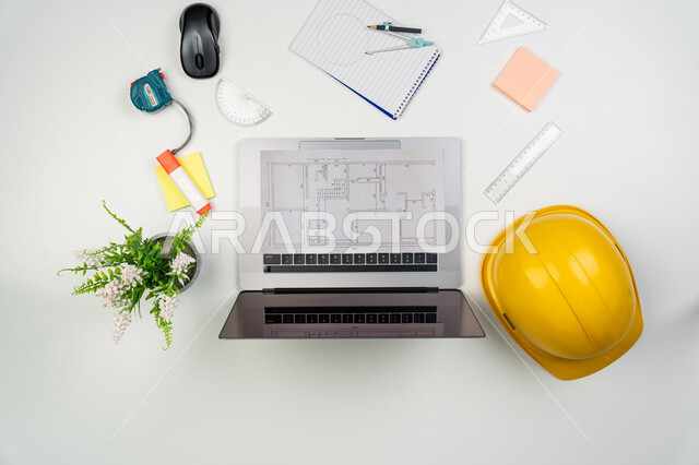 المعدات والأدوات ومخططات البناء على طاولة مكتبية، مهن ووظائف هندسية، صورة علوية عامودية لخوذة الوقاية البيضاء واللابتوب على المكتب، متابعة الأعمال والتصاميم عبر جهاز الحاسوب المحمول، إنجاز مهام العمل ورسم المشاريع بتقنيات حديثة، خلفية بيضاء