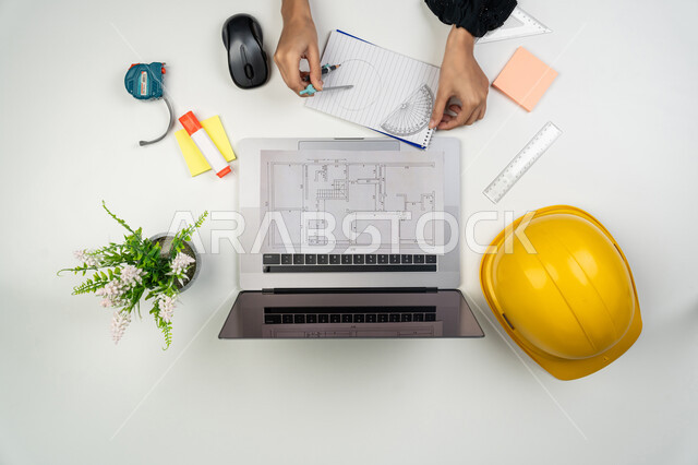 مراجعة المشاريع وتدقيقها، أدوات هندسية لإكمال رسم مخطط البناء، الإنشاءات المعمارية والهندسة، متابعة استراتيجات المشروع, بورتريه علوي عامودي ليدي مهندسة عربية خليجية اماراتية تعمل على اللابتوب، امرأة سعودية امام مكتب عليه خوذة الحماية والمخططات، خلفية بيضاء