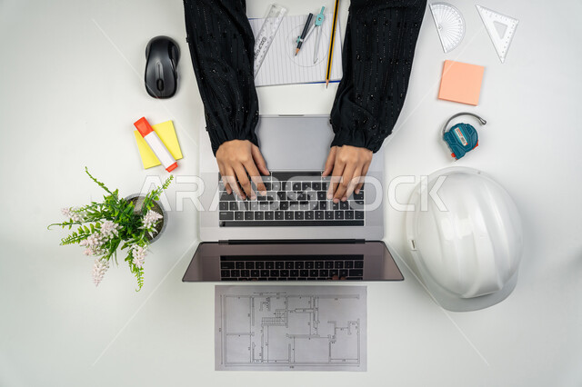 الإنشاءات المعمارية والهندسة، متابعة استراتيجات المشروع, بورتريه علوي عامودي ليدي مهندسة عربية خليجية اماراتية تعمل على اللابتوب، امرأة سعودية امام مكتب عليه خوذة الحماية والمخططات، أدوات هندسية لإكمال رسم مخطط البناء، تدقيق المشاريع ومراجعتها، خلفية بيضاء