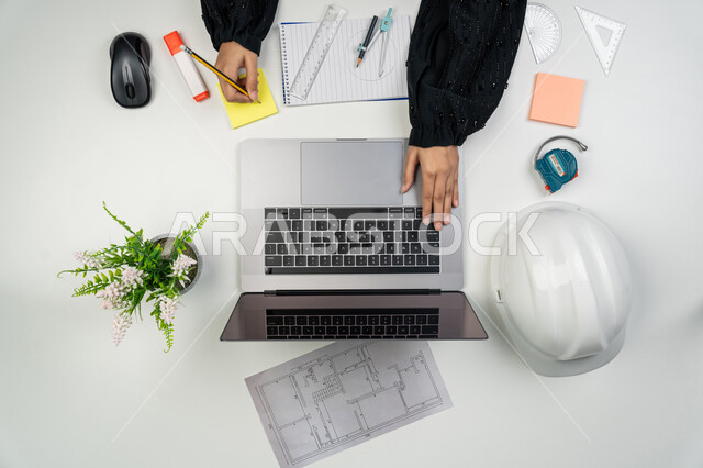 الهندسة والإنشاءات المعمارية، متابعة استراتيجات المشروع, بورتريه علوي عامودي ليدي مهندسة عربية خليجية اماراتية تعمل على اللابتوب، امرأة سعودية امام مكتب عليه خوذة الحماية والمخططات، أدوات هندسية لإكمال رسم مخطط البناء، تدقيق المشاريع ومراجعتها، خلفية بيضاء