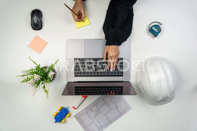 Project strategy follow-up, architectural engineering and construction, vertical top portrait of an Arab Gulf Emirati female engineer working on laptop, Saudi woman in front of desk with safety helmet and blueprints, engineering tools to complete building plan drawing, project auditing and review, white background