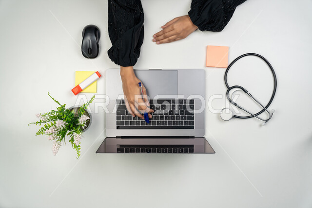 تقدم القطاع الصحي، اعطاء الاستشارات للمرضى عن بعد عبر الحاسوب، مفهوم الرعاية الصحية، دمج التقنيات الحديثة بالمجالات الطبية, بورتريه علوي عامودي ليدي طبيبة عربية خليجية اماراتية تستخدم اللابتوب بجانبها سماعات طبية، سيدة سعودية تعمل في مجال الطب، خلفية بيضاء