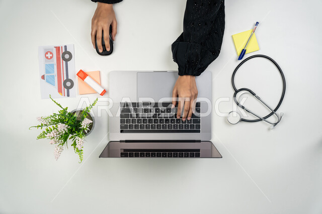 Providing remote consultations to patients via computer, development of the health sector, integration of modern technologies in medical fields, vertical upper portrait of an Arab Gulf Emirati female doctor using a laptop next to her with medical stethoscopes, a Saudi woman working in the field of medicine, the concept of health care, white background