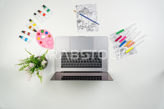 التدريب والتعلم من خلال المواقع الإلكترونية، اقتناء الألوان المائية والخشبية ومستلزمات الرسم أونلاين، الشراء والبيع عبر الانترنت، صورة علوية عامودية لجهاز الحاسوب المحمول مع أوراق ومجموعة من أقلام التلوين وكوب من القهوة، خلفية بيضاء