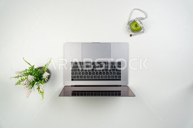 الالتزام في برنامج صحي متكامل من خلال البحث على المواقع الإلكترونية، التسوق لشراء المستلزمات الرياضية، صورة علوية عامودية لجهاز الحاسوب المحمول مع تفاحة ومتر القياس، الحفاظ على اللياقة والصحة البدنية، خلفيه بيضاء