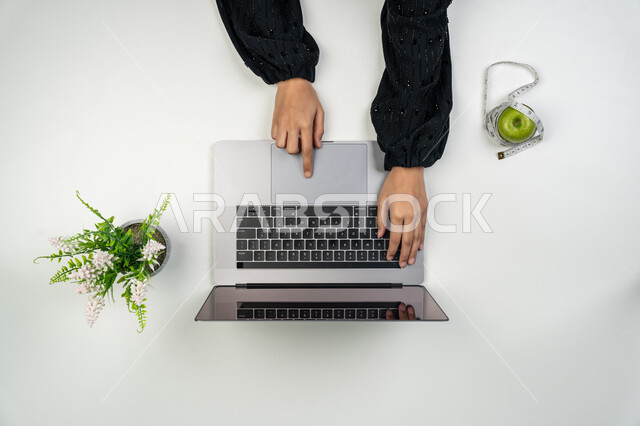 التسوق لشراء المستلزمات الرياضية، البحث عن برنامج صحي متكامل عبر المواقع الإلكترونية، المحافظة على اللياقة والصحة البدنية، بورتريه علوي عامودي ليدي إمرأة عربية خليجية اماراتية تستخدم اللابتوب، سيدة سعودية بجانبها تفاحة مع متر القياس، خلفيه بيضاء