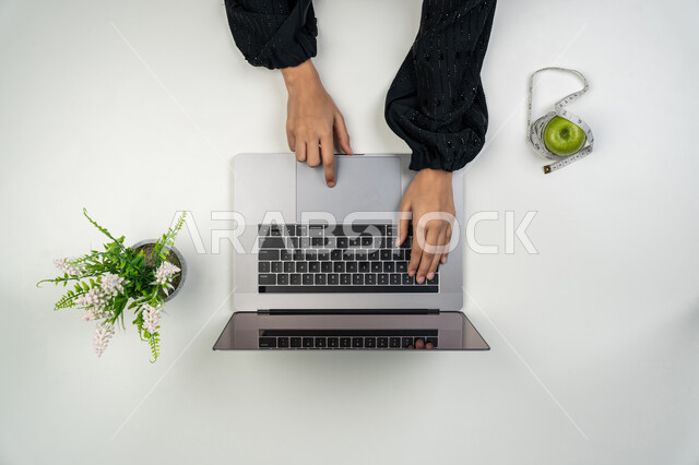 Searching for a comprehensive health program through websites, maintaining fitness and physical health, shopping for sports supplies, vertical upper portrait of an Emirati Gulf Arab woman using a laptop, a Saudi woman with an apple next to her with a measuring tape, white background