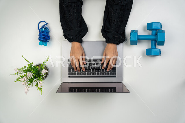 البحث عن برنامج صحي متكامل عبر المواقع الإلكترونية، التسوق لشراء المستلزمات الرياضية، بورتريه علوي عامودي ليدي إمرأة عربية خليجية اماراتية تستخدم اللابتوب، سيدة سعودية بجانبها الأثقال (الدمبلز) وحبل النط ، الحفاظ على اللياقة والصحة البدنية، خلفيه بيضاء