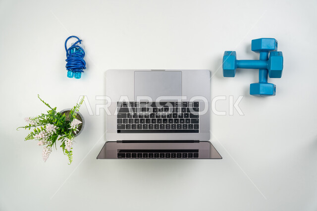 الالتزام  في برنامج صحي متكامل من خلال البحث على المواقع الإلكترونية، التسوق لشراء المستلزمات الرياضية، صورة علوية عامودية لجهاز الحاسوب المحمول مع الأثقال (الدمبلز) وحبل النط ، الحفاظ على اللياقة والصحة البدنية، خلفيه بيضاء
