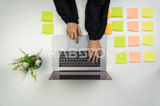 استخدام الأجهزة التقنية الحديثة في الأعمال المنزلية، تثبيت أوراق ملاحظات تذكارية ملونة، تسجيل الملاحظات ووتدوين الأفكار، بورتريه علوي عامودي لايدي امرأة عربية خليجية اماراتية تعمل على الحاسوب المحمول، شابة سعودية تقوم بانجاز مهام العمل عن بعد، خلفية بيضاء