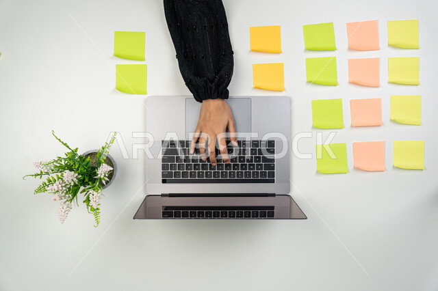 تثبيت أوراق ملاحظات تذكارية ملونة، استخدام الأجهزة التقنية الحديثة في الأعمال المنزلية، تسجيل الملاحظات ووتدوين الأفكار، بورتريه علوي عامودي لايدي امرأة عربية خليجية اماراتية تعمل على الحاسوب المحمول، شابة سعودية تقوم بانجاز مهام العمل عن بعد، خلفية بيضاء
