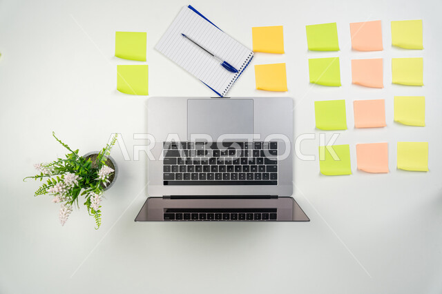 دفتر ملاحظات وأوراق وأقلام ملونة، استخدام الأجهزة التقنية الحديثة والمتطورة في الأعمال المنزلية، انجاز مهام العمل عن بعد، تسجيل الملاحظات ووتدوين الأفكار، صورة علوية عامودية لحاسوب محمول ومجموعة من القرطاسية على طاولة مكتبية، خلفية بيضاء