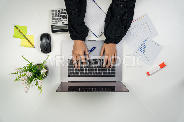 إنخفاض وارتفاع نسبة الأسهم، حساب نسبة الأرباح المالية، بورتريه علوي عامودي لأيدي امرأة عربية خليجية اماراتية تعمل على جهاز الحاسوب المحمول، شابة سعودية تدون الملاحظات وتستخدم الألة الحاسبة، مهن ووظائف نسائية، متابعة مخطط الرسم البياني للنمو الاقتصادي، خلفية بيضاء