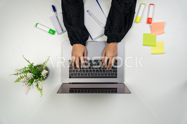 القيام بانجاز مهام العمل عن بعد، تسجيل الملاحظات والأفكار، بورتريه علوي عامودي ليدي امرأة عربية خليجية اماراتية تقوم بالكتابة على دفتر وبجانبها أوراق وأقلام ملونة، سيدة سعودية تستخدم جهاز الحاسوب، استخدام الأجهزة التقنية الحديثة في الأعمال المنزلية، خلفية بيضاء